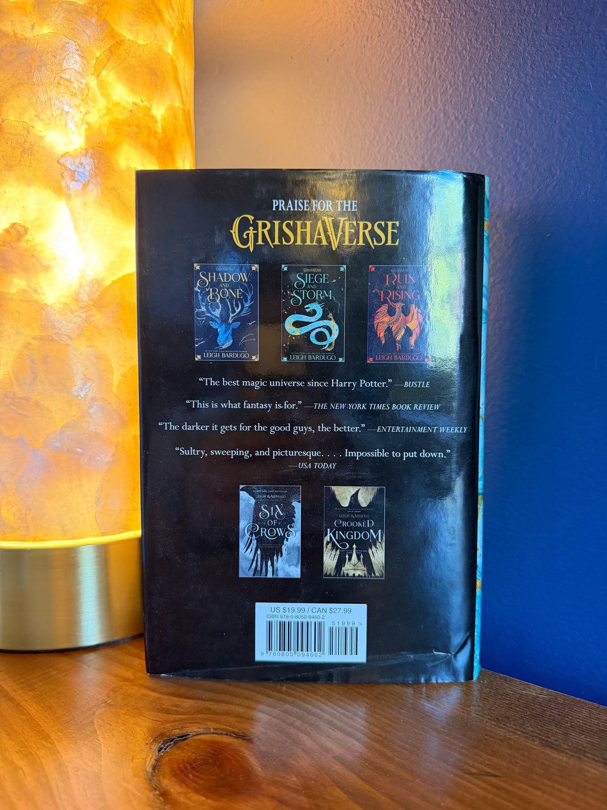 The back cover of a book by Leigh Bardugo is shown standing upright on a wooden surface, with a glowing orange lamp on the left and a blue wall in the background.