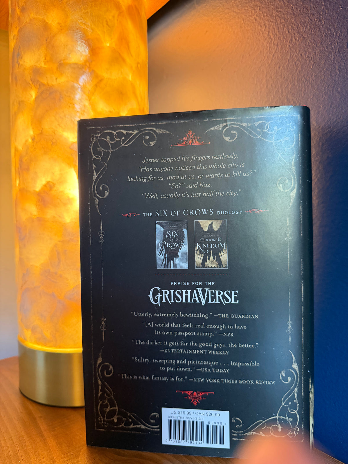 The back cover of a book by Leigh Bardugo, displayed on a wooden surface next to a glowing orange lamp.