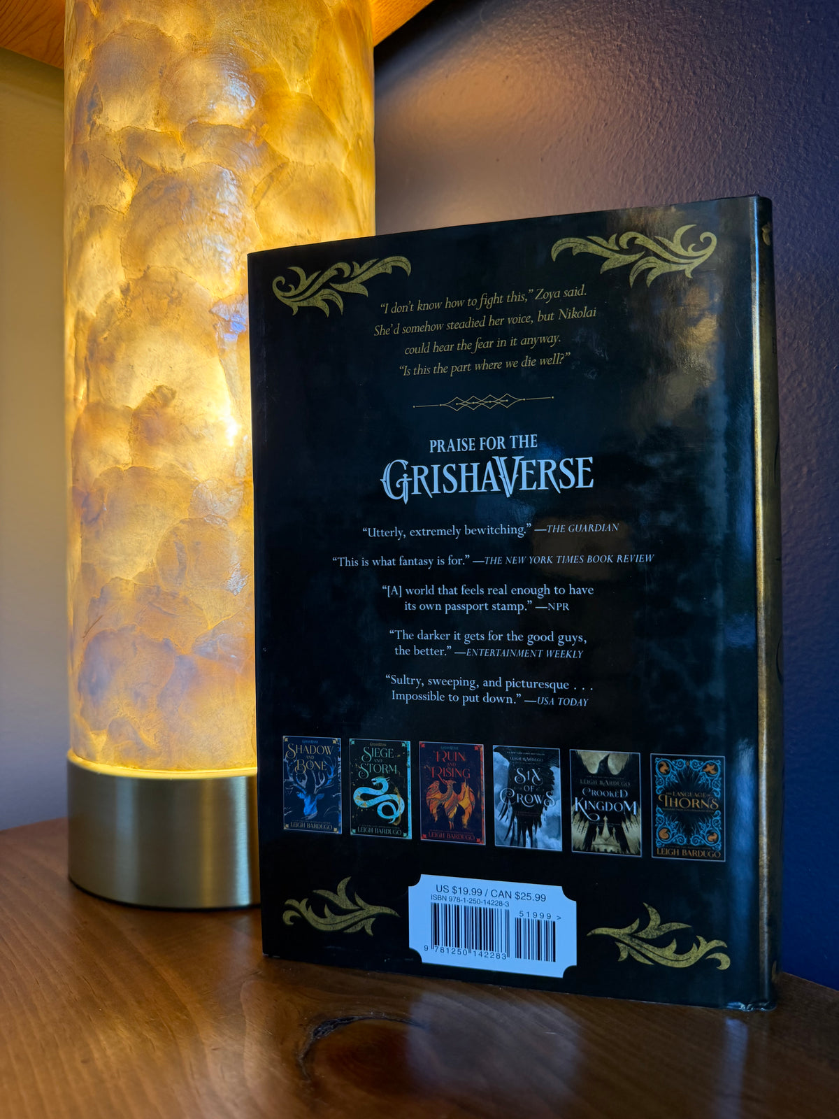 The back cover of a book by Leigh Bardugo is shown standing upright on a wooden table next to a glowing cylindrical lamp. The cover is dark with gold decorative flourishes and several small book cover thumbnails along the bottom.