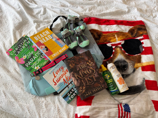 A summer beach flat-lay on a white blanket with a stack of romance paperbacks, including Beach Read by Emily Henry and Every Summer After by Carley Fortune, next to a striped beach towel, sunscreen, and a pair of dog booties.