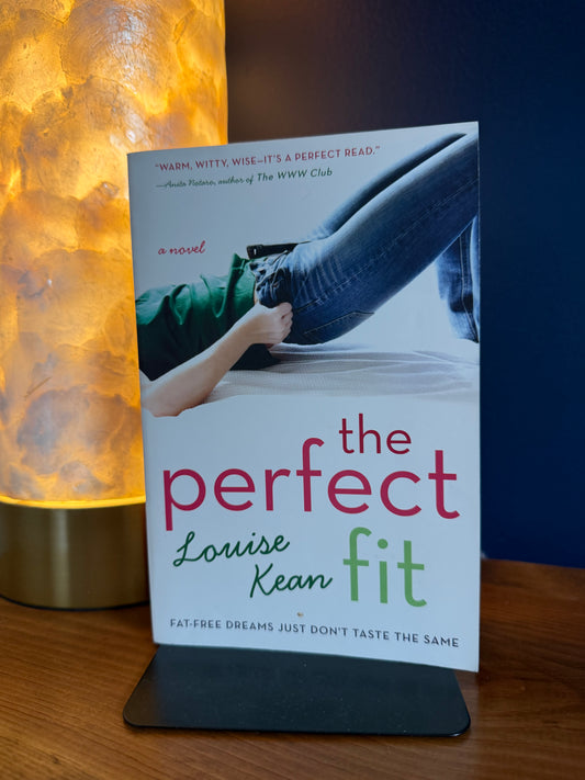 Book titled 'The Perfect Fit' by Louise Kean on a stand with a warm light source in the background.