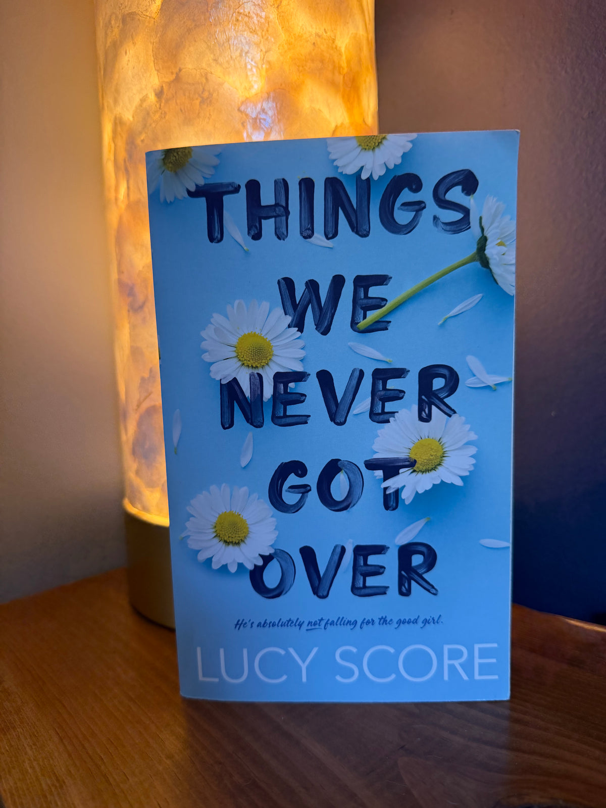 The book cover of 'Things We Never Got Over' by Lucy Score, featuring a light blue background with white daisies and bold dark blue text, displayed on a wooden surface in front of a glowing lamp.