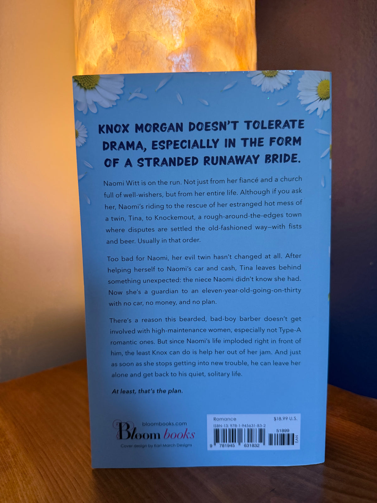 The back cover of a paperback book by Bloom Books, featuring a light blue background with white daisies and black text, photographed on a wooden surface with warm lighting.