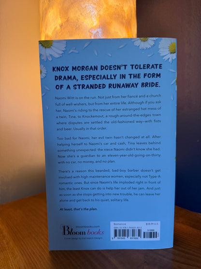 The back cover of a paperback book by Bloom Books, featuring a light blue background with white daisies and black text, photographed on a wooden surface with warm lighting.
