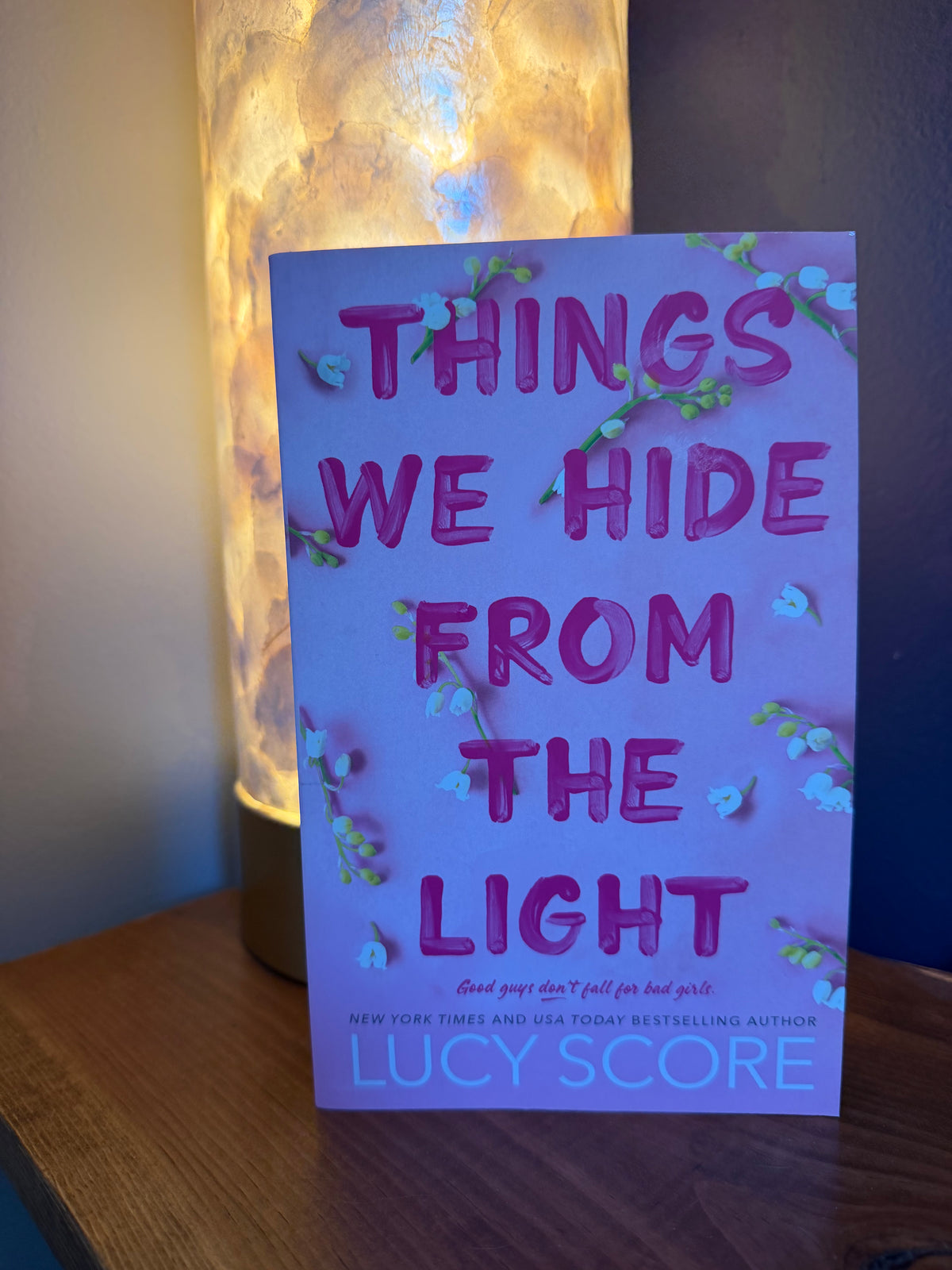 The book 'Things We Hide From the Light' by Lucy Score, featuring a light pink cover with pink title text and small white flowers, standing on a wooden surface next to a glowing lamp.