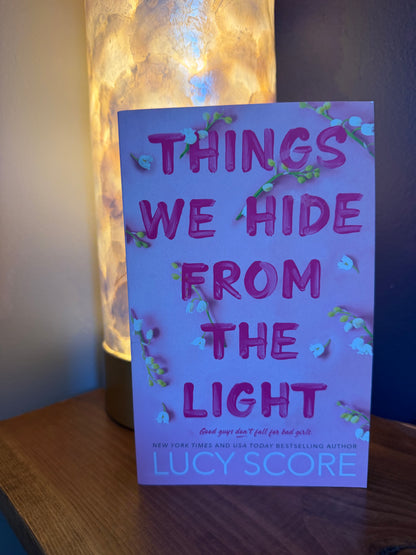 The book 'Things We Hide From the Light' by Lucy Score, featuring a light pink cover with pink title text and small white flowers, standing on a wooden surface next to a glowing lamp.
