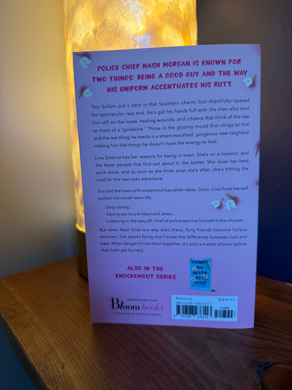 The back cover of a book, published by Bloom Books, displayed on a wooden surface with a warm light shining from behind.