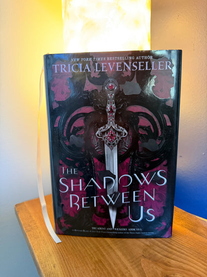 The hardcover book 'The Shadows Between Us' by Tricia Levenseller, featuring a fantasy cover with a silver dagger and purple shadows, resting on a wooden table next to a glowing lamp.