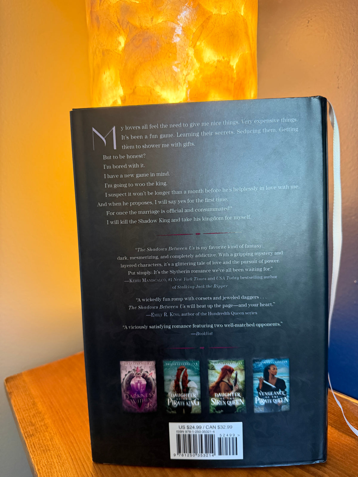 The back cover of a book titled ''The Shadows Between Us' by Tricia Levenseller is shown standing upright on a wooden surface, with glowing orange light in the background.