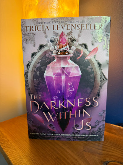 The book cover of 'The Darkness Within Us' by Tricia Levenseller, featuring a glowing purple crystal bottle, displayed on a wooden surface with a warm orange lamp in the background.