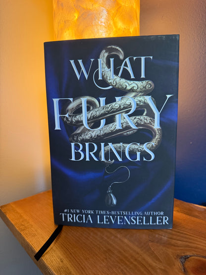 The book cover of 'What Fury Brings' by Tricia Levenseller, featuring a dark blue background with a silver serpent and light blue title text, displayed on a wooden surface with a warm lamp in the background.