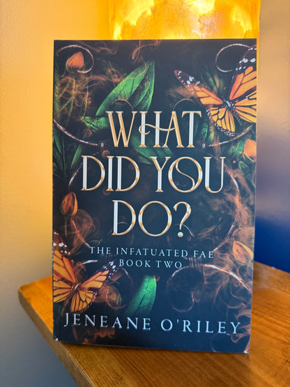 A book titled "What Did You Do?" by Jeneane O'Riley, featuring a dark cover with orange monarch butterflies, green leaves, and swirling smoke, displayed on a wooden surface with a warm glowing lamp in the background.