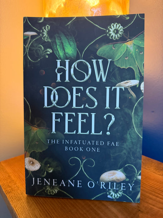 The book 'How Does It Feel? The Infatuated Fae Book One' by Jeneane O'Riley, standing upright on a wooden surface with a warm, glowing background.