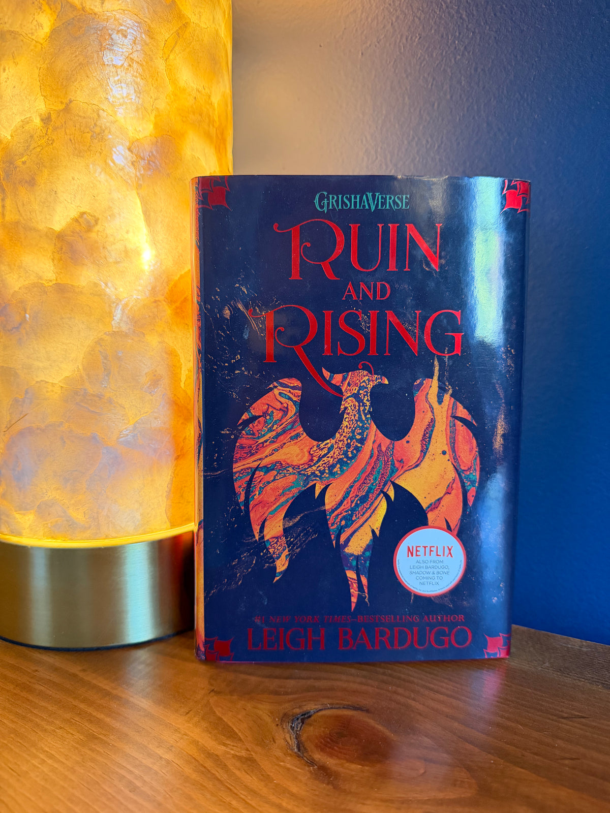 The book 'Ruin and Rising' by Leigh Bardugo, published under the GrishaVerse series, is displayed upright on a wooden surface next to a glowing yellow lamp. The book cover features a black background with a stylized illustration of a bird in orange, red, and gold patterns, and a prominent red title.
