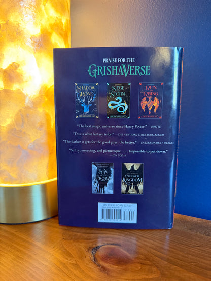 The back cover of a book by Leigh Bardugo is shown standing upright on a wooden shelf, next to a glowing warm lamp and a dark blue wall.