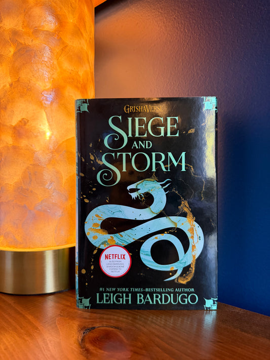 The book 'Siege and Storm' by Leigh Bardugo, part of the Grishaverse series, is standing on a wooden shelf next to a glowing orange lamp. The book cover is black with a light blue coiled snake and gold accents, featuring a red Netflix badge.