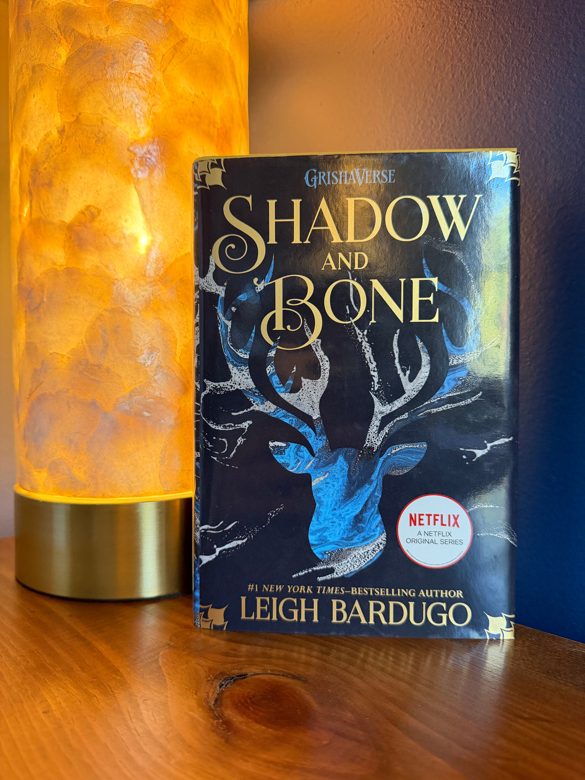 The book 'Shadow and Bone' by Leigh Bardugo is displayed upright on a wooden shelf next to a glowing orange lamp. The book cover features a dark blue and black background with a silver stag illustration and gold lettering.