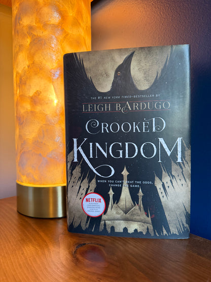 A copy of the book 'Crooked Kingdom' by Leigh Bardugo stands upright on a wooden surface next to a glowing orange lamp. The book cover features a dark illustration of a raven and the title in large white lettering, with a Netflix badge near the bottom.