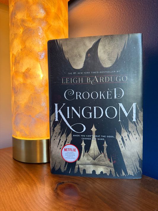 A copy of the book 'Crooked Kingdom' by Leigh Bardugo stands upright on a wooden surface next to a glowing orange lamp. The book cover features a dark illustration of a raven and the title in large white lettering, with a Netflix badge near the bottom.