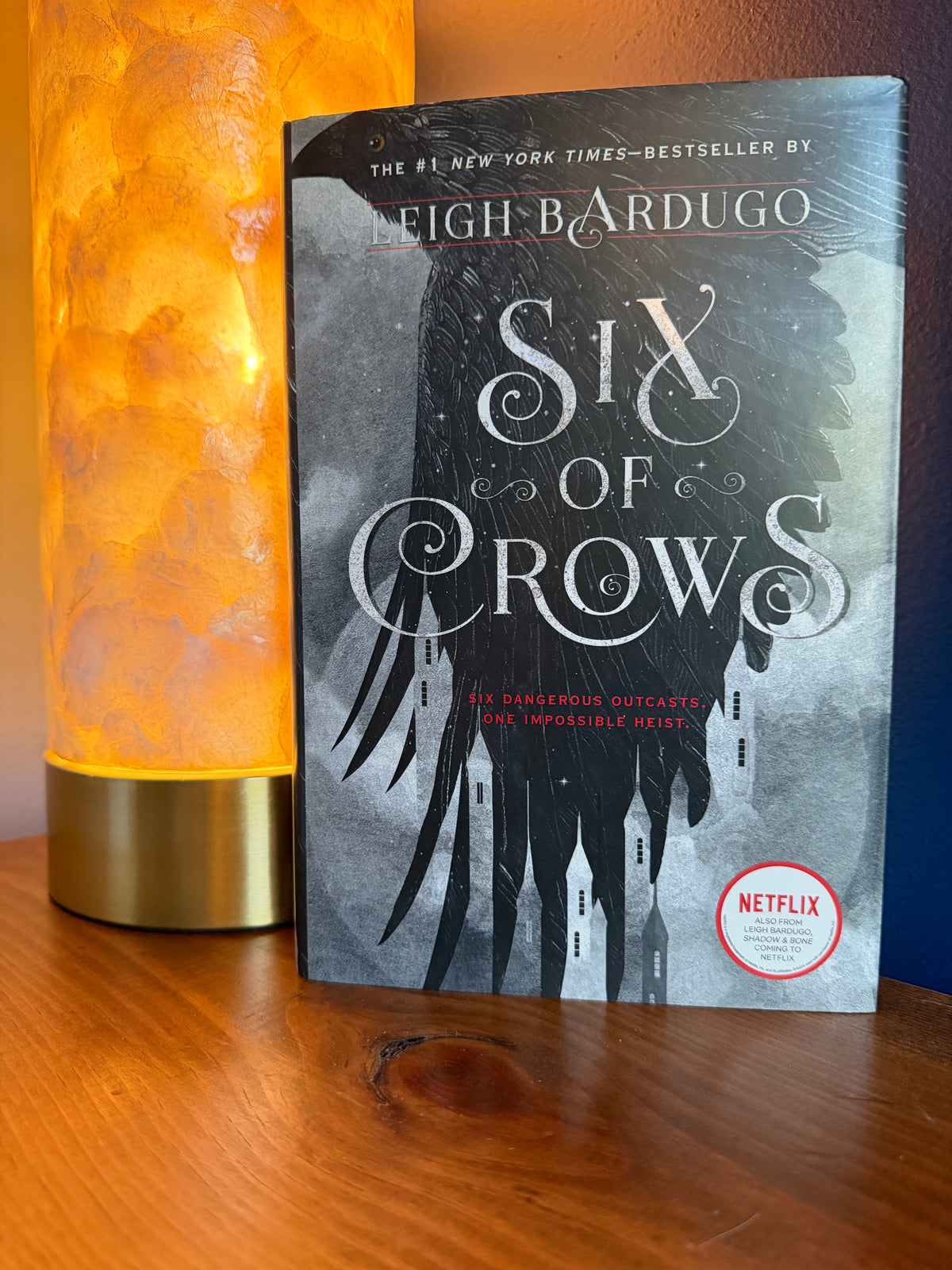 Six of Crows book by Leigh Bardugo on a wooden shelf next to a glowing orange lamp, with a Netflix badge on the cover.