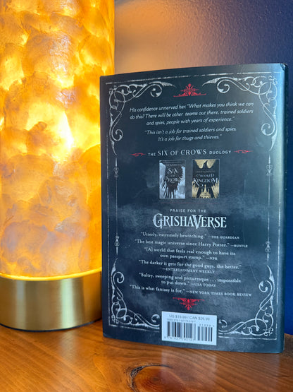 The back cover of a book by Leigh Bardugo, displayed on a wooden surface next to a glowing warm lamp.