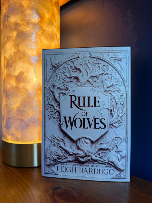 The book 'Rule of Wolves' by Leigh Bardugo stands upright on a wooden surface next to a glowing, textured cylindrical lamp. The book cover is a pale, textured gray with embossed wolf graphics and the title in black lettering.
