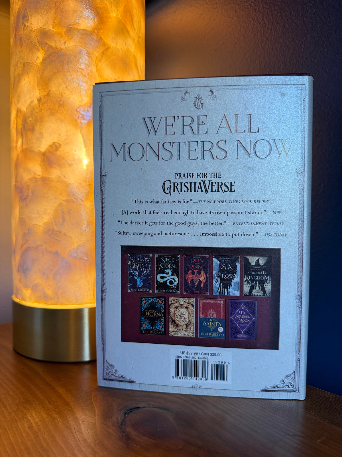 The back cover of a book by Leigh Bardugo, displayed on a wooden surface next to a glowing orange lamp.