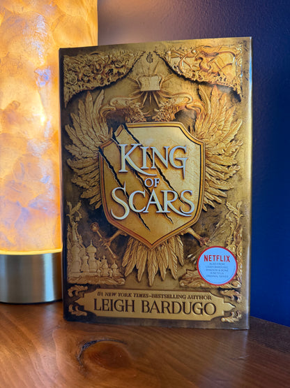 King of Scars book by Leigh Bardugo on a wooden table next to a glowing lamp, with a blue wall in the background. A round Netflix sticker is visible on the front cover.